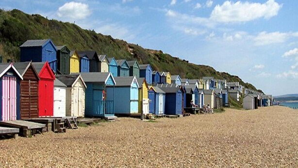 A row of colorful beach huts along a pebbled beach with a grassy hill in the background. - Auplands (Lymington)