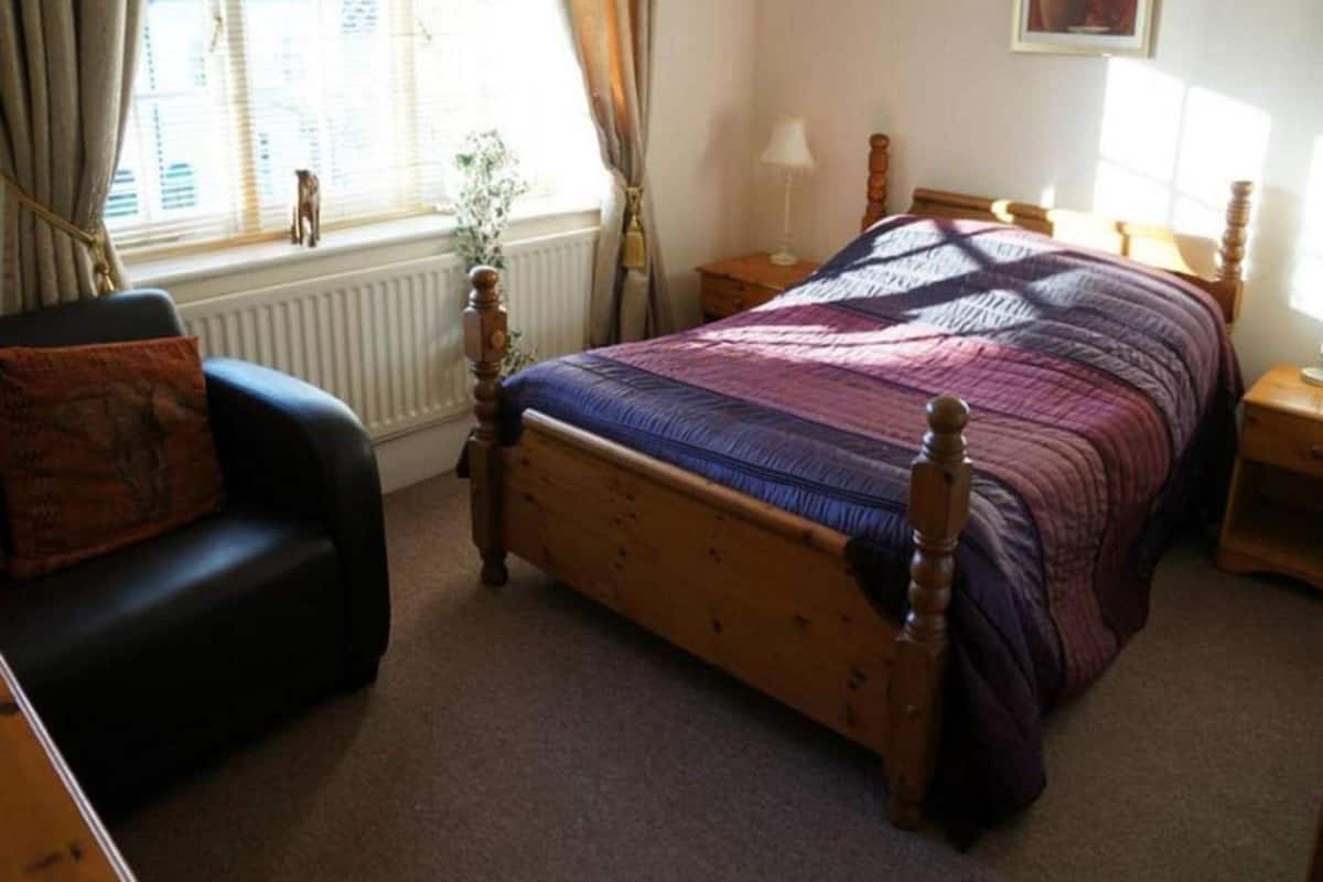 A bedroom with a wooden bed, a black armchair, a bedside table, and a window with curtains.