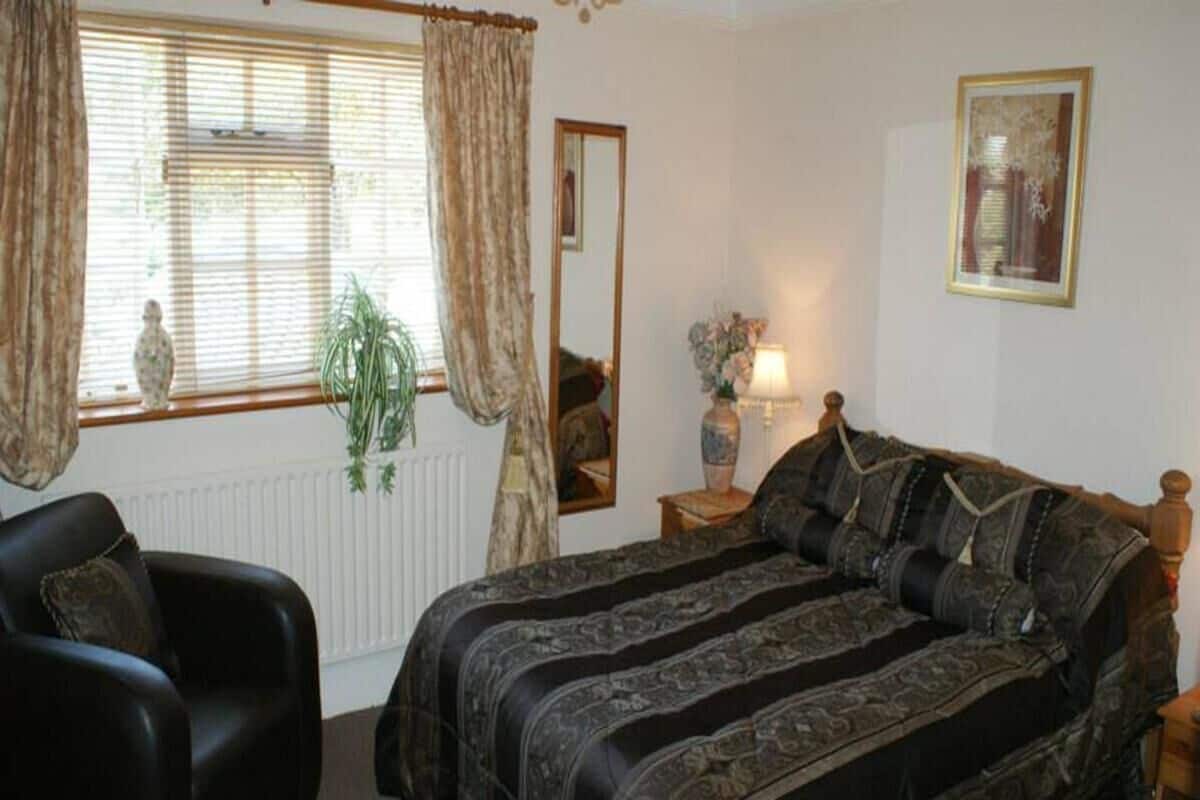 A bedroom with a large bed, a chair, a bedside table with a lamp, a mirror, and a window with curtains.