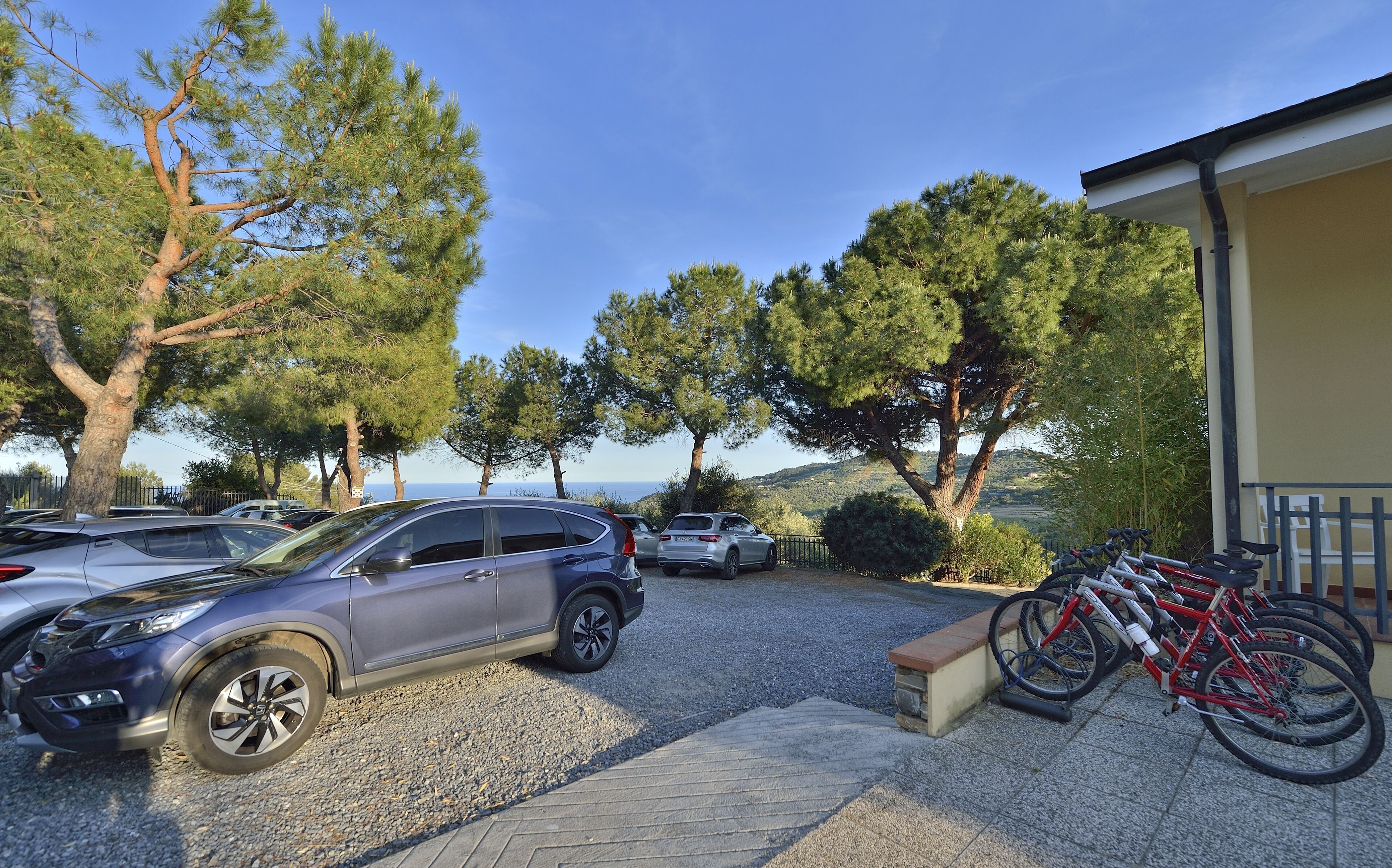 A parking area with cars and bicycles, surrounded by trees and a building with a beige facade.