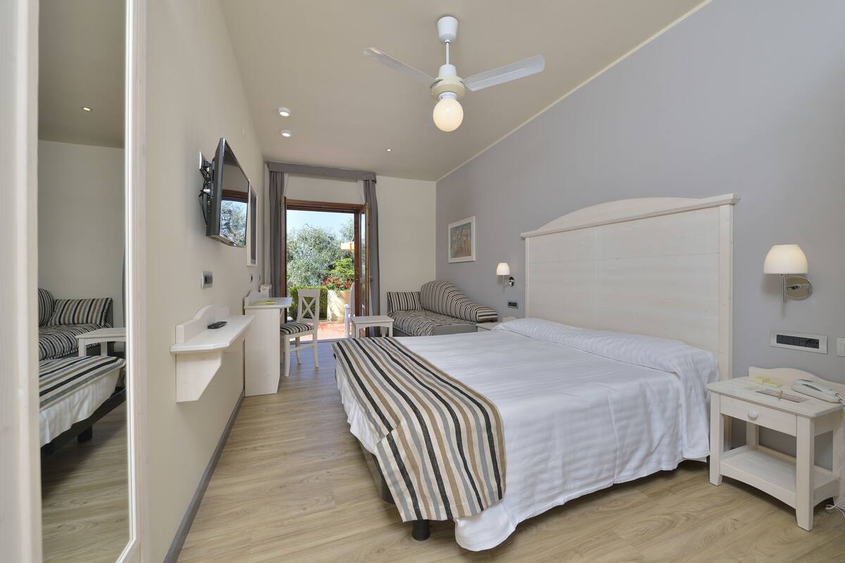 A bedroom with a large bed, a bedside table, a nightstand, a ceiling fan, a mirror, and a window with curtains.