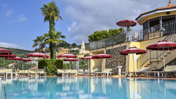 A swimming pool with sun umbrellas and lounge chairs, a stone staircase, and a view of a hill with buildings. - Hotel Liliana Diano Marina (Diano Marina)