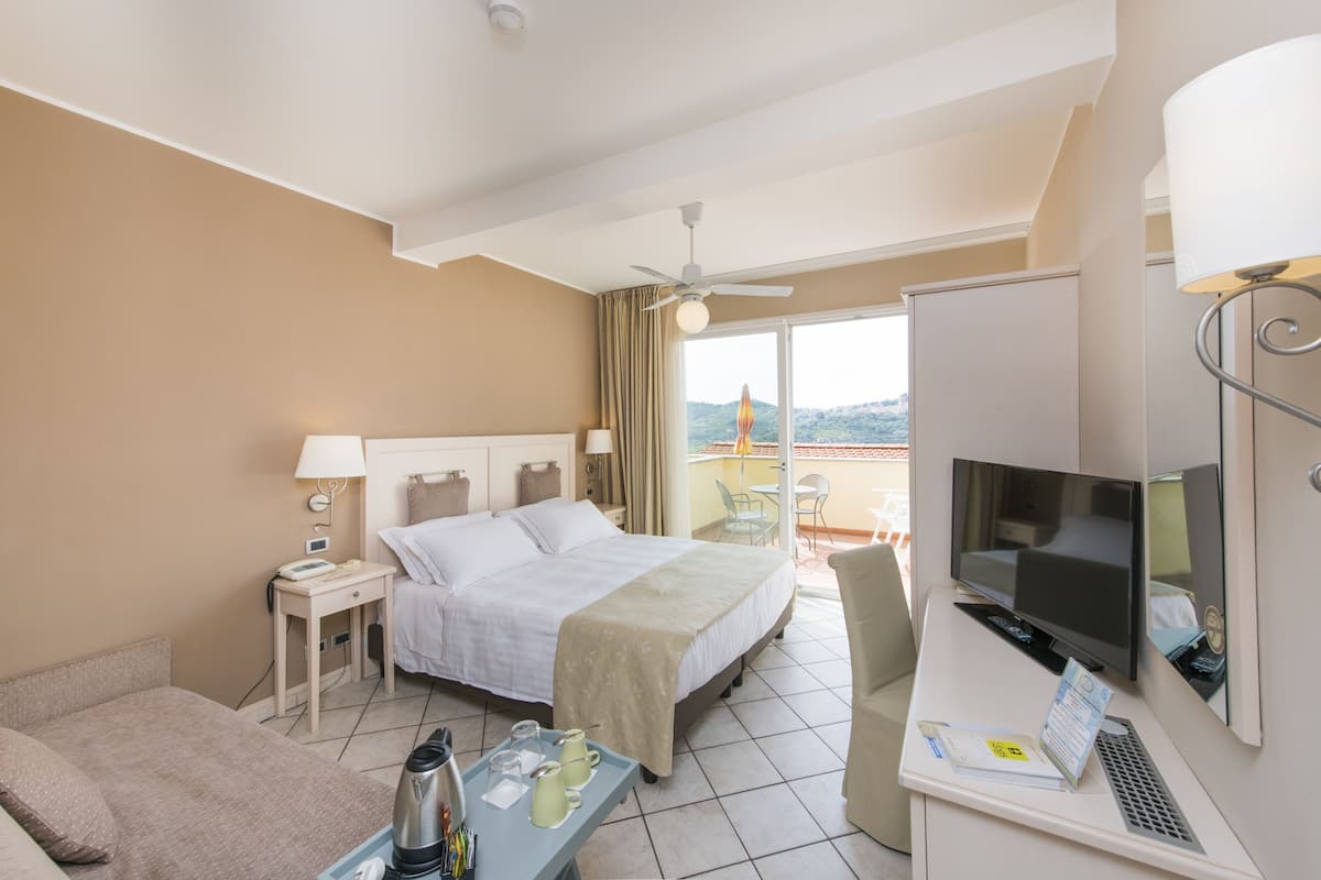 A hotel room with a bed, a desk with a TV, a chair, a small table with a kettle and cups, and a balcony with a view.
