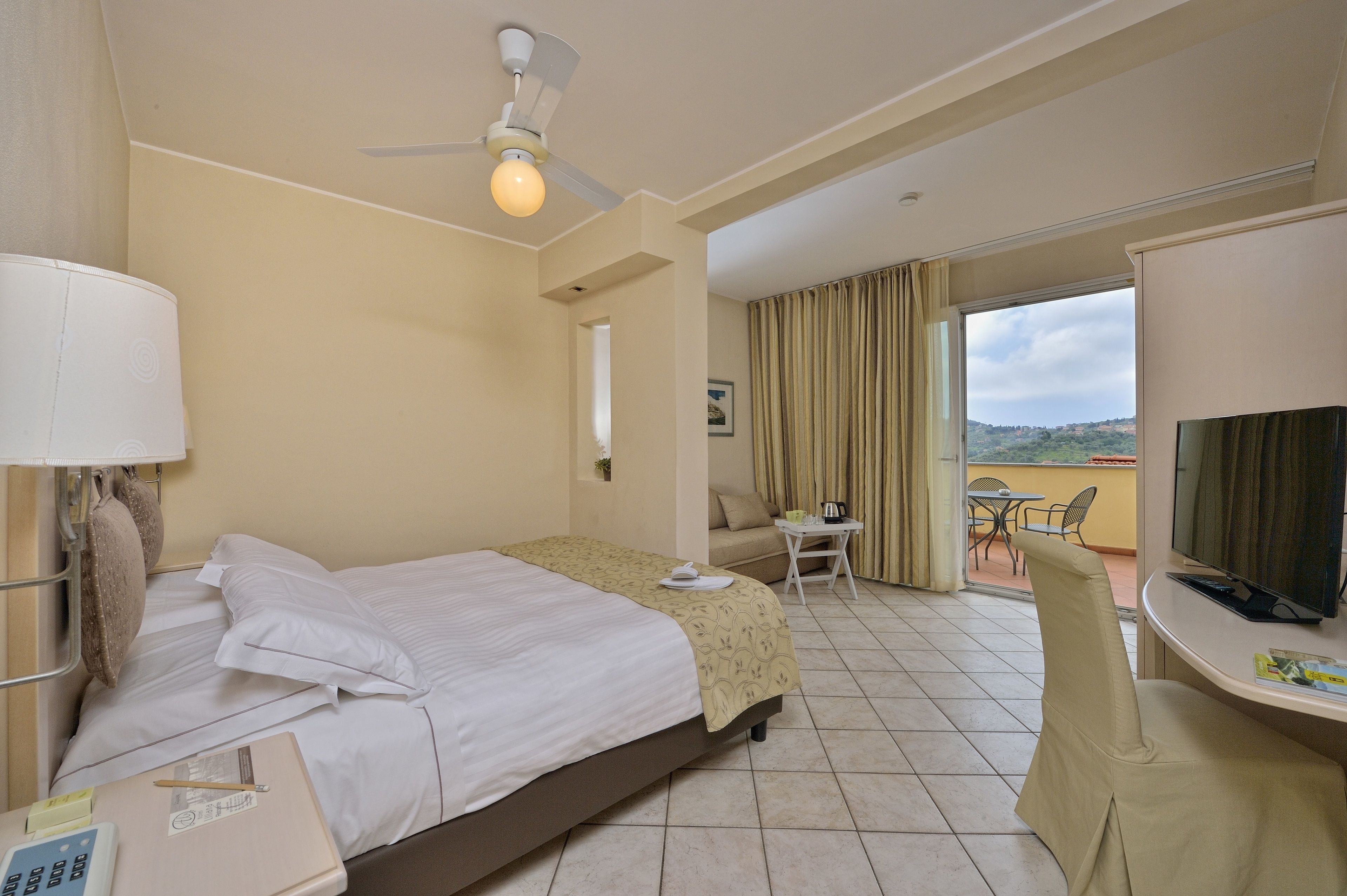 A hotel room with a bed, a desk with a chair, a television, and a balcony with a table and chairs.