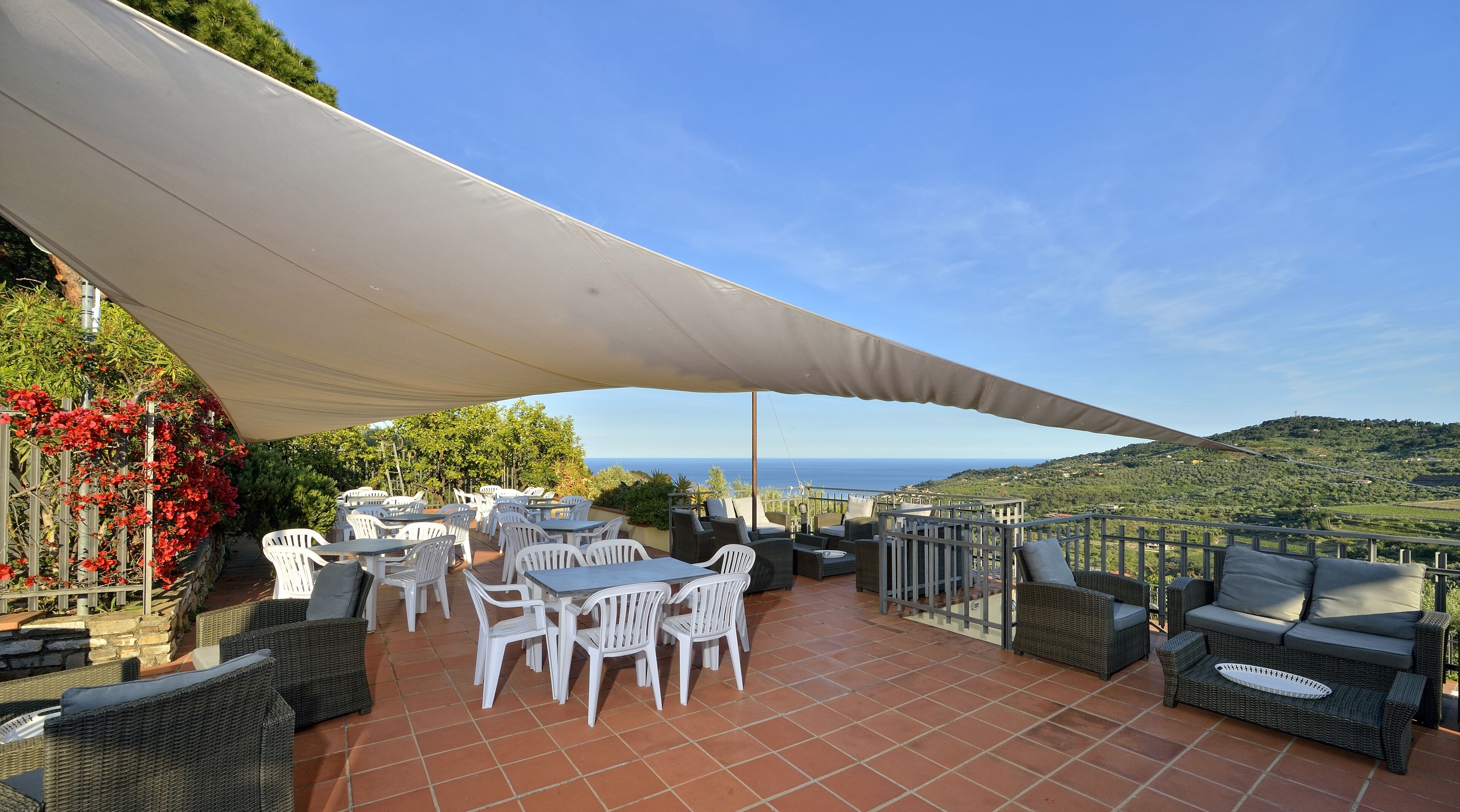 Un patio al aire libre con sillas y mesas de plástico blanco, un toldo grande y una vista panorámica del mar y las colinas.