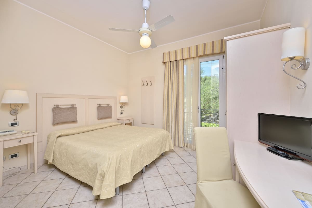 A hotel room with a bed, a desk with a television, a chair, a ceiling fan, and a window with curtains.