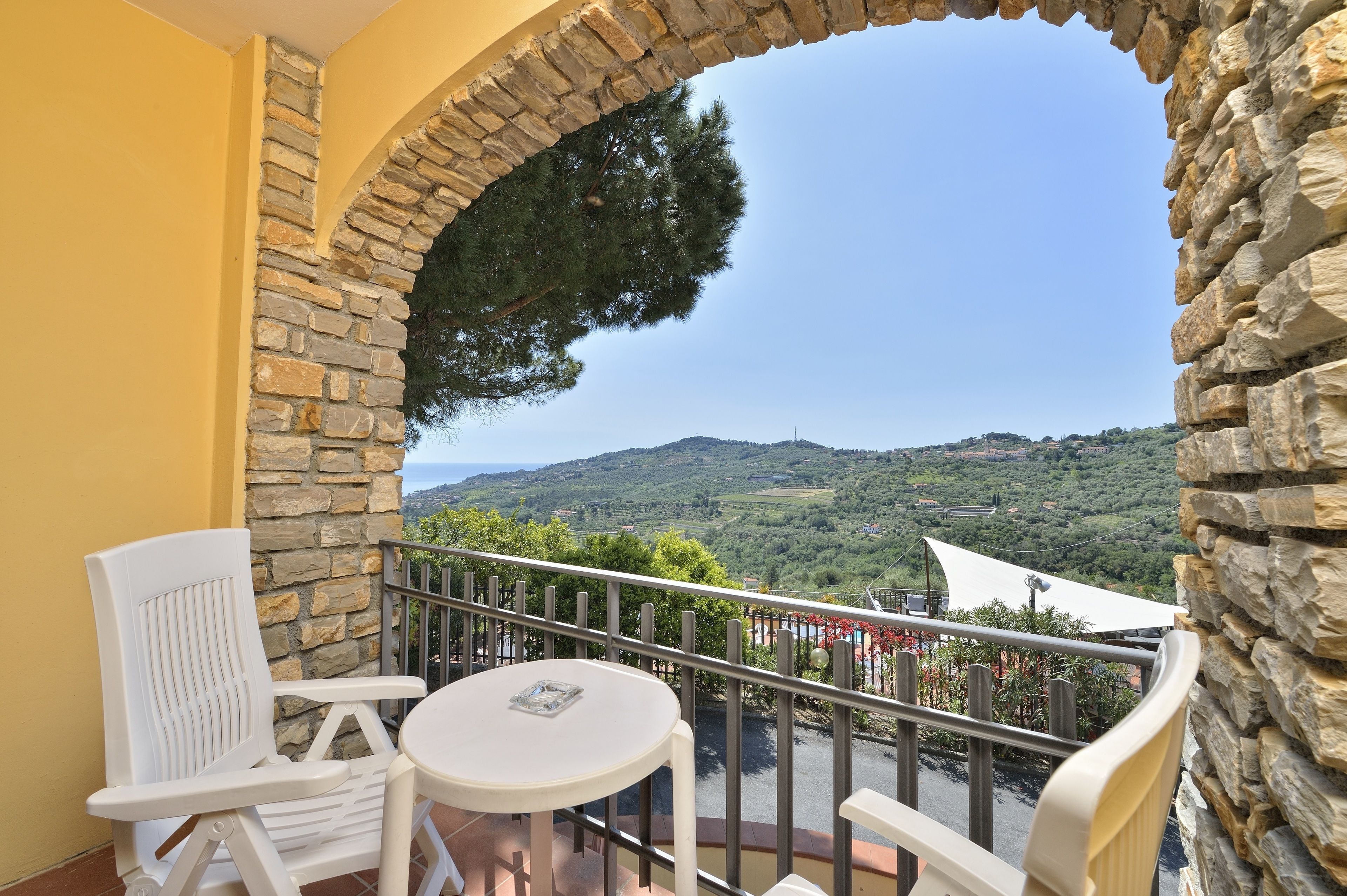 Un balcone con pareti in pietra, una sedia bianca, un tavolino e vista su un paesaggio verde con colline.