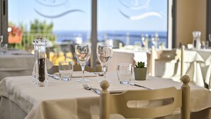 A dining table set with wine glasses, a salt and pepper shaker, and a small plant, with a view of the sea through large windows. - Hotel Liliana Diano Marina (Diano Marina)