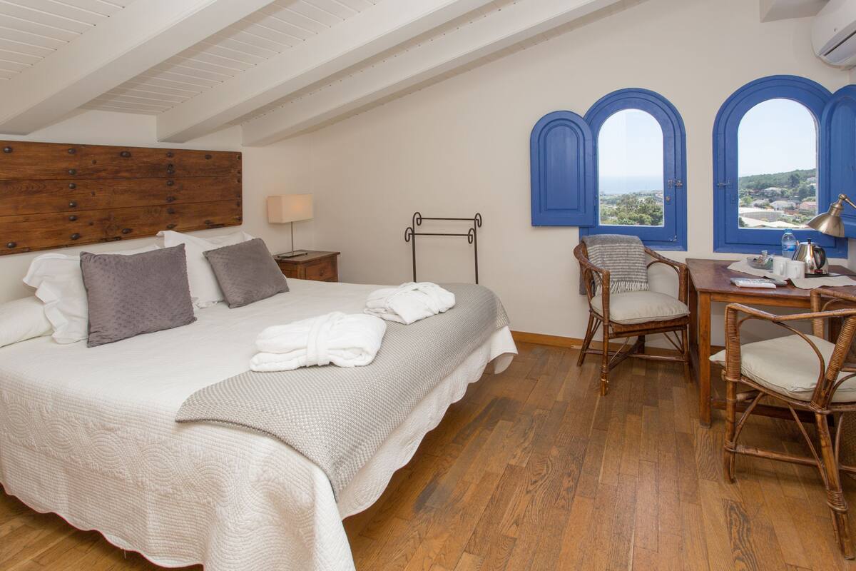 Ein Schlafzimmer mit einem großen Bett, einem Holzkopfende, Holzboden, einem Stuhl, einem Tisch und Meerblick durch blaue Fensterrahmen.