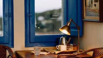 Una habitación con una mesa de madera, dos sillas de mimbre, una lámpara de latón, una cafetera y un cuadro en la pared.