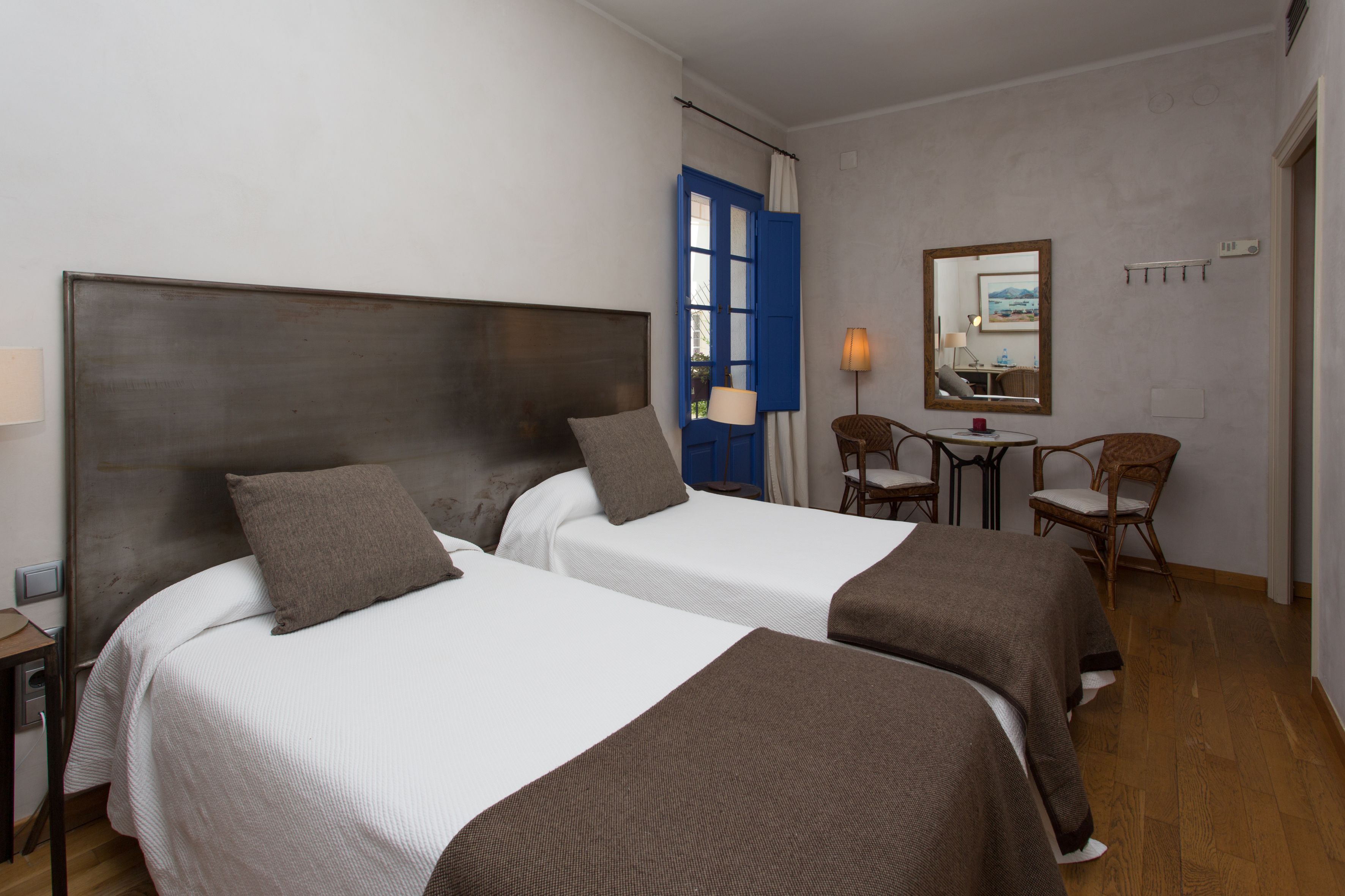 A hotel room with two beds, a small table with a lamp, two chairs, and a mirror on the wall.