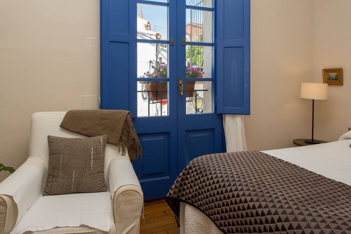 Ein Zimmer mit einer blauen Tür, einem weißen Sessel mit einer braunen Decke, einer gemusterten Bettdecke und einem kleinen Tisch mit einer Lampe.