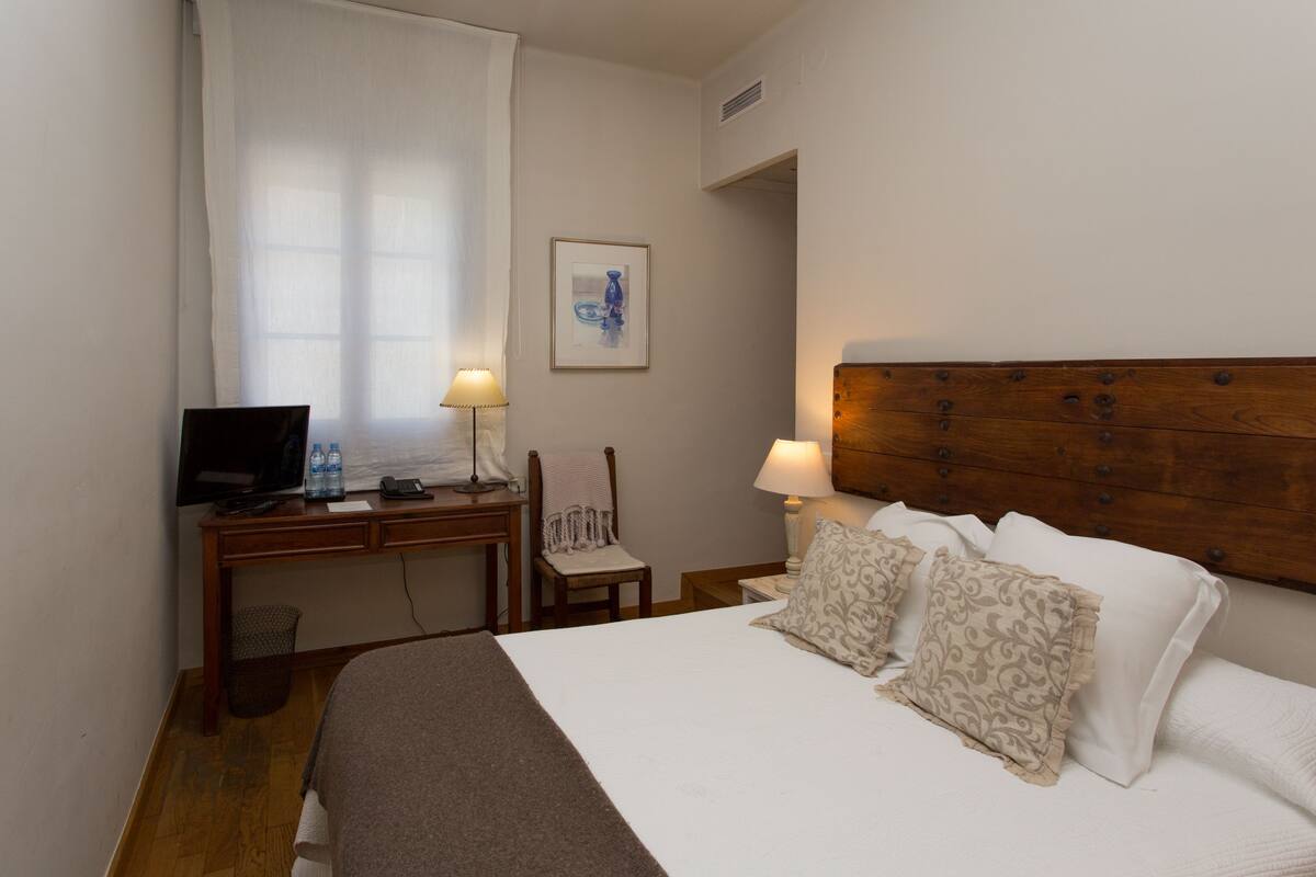 Ein Hotelzimmer mit einem Bett, einem Schreibtisch mit Computer und Stuhl, einer Lampe und einem gerahmten Bild an der Wand.