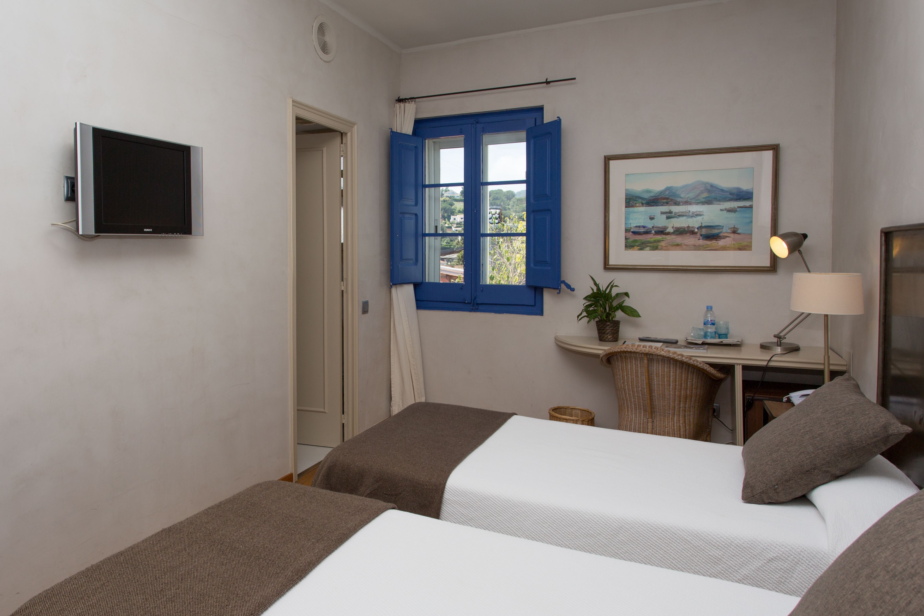 Une chambre d’hôtel avec deux lits, un bureau avec une lampe, une télévision, une fenêtre avec des volets bleus et un tableau au mur.