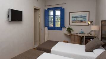 Une chambre d’hôtel avec deux lits, un bureau avec une lampe, une télévision, une fenêtre avec des volets bleus et un tableau au mur.