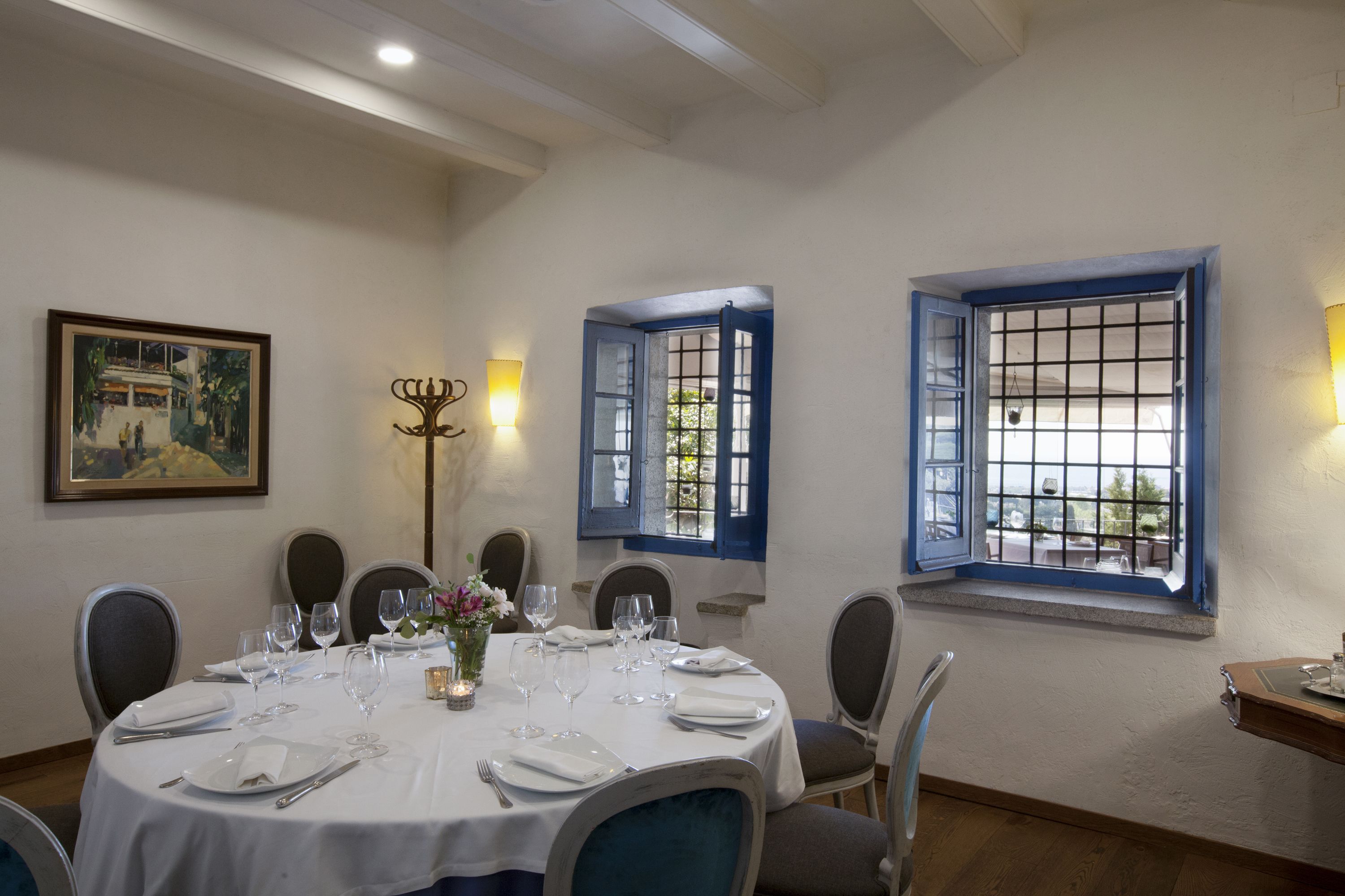 Une salle à manger avec une table ronde dressée pour quatre personnes, des nappes blanches et des volets bleus aux fenêtres.