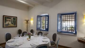 Une salle à manger avec une table ronde dressée pour quatre personnes, des nappes blanches et des volets bleus aux fenêtres.