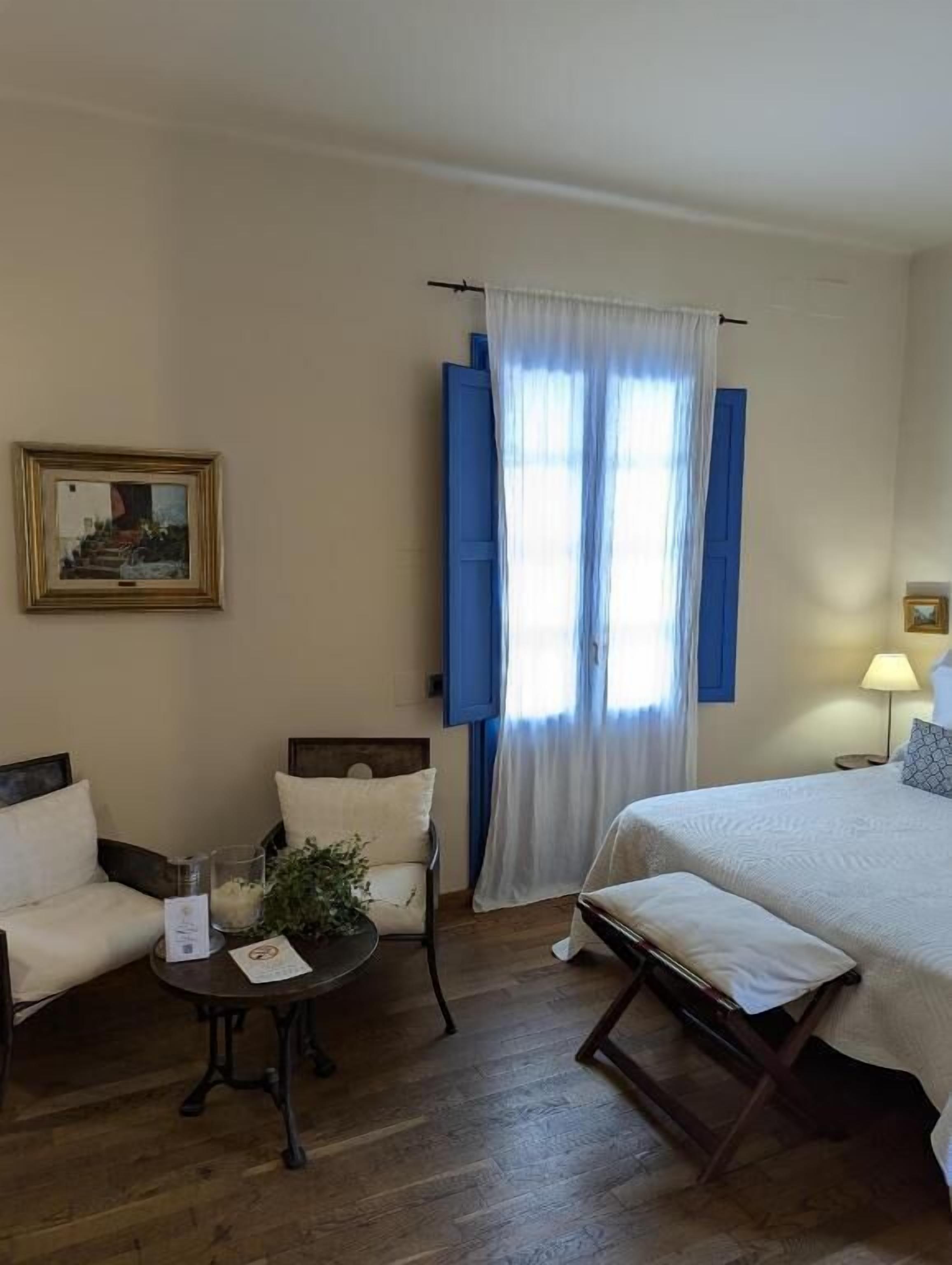 A hotel room with a bed, a chair, a small table with a plant, a lamp, and a framed picture on the wall.