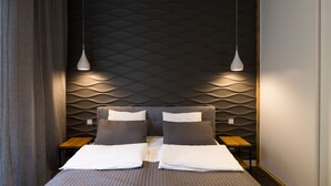 Une chambre d’hôtel moderne avec un mur texturé, deux lits avec une literie grise, une table de chevet en bois et une lampe suspendue.
