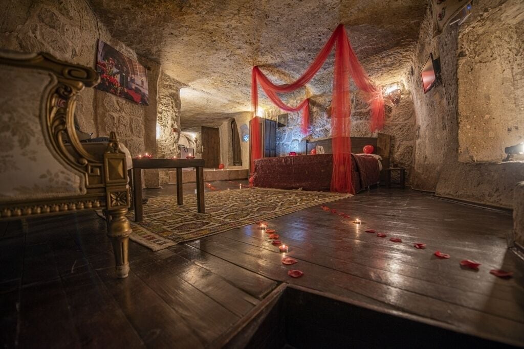 Una habitación con suelo de madera, cama con cortinas rojas, un espejo y una pequeña mesa con velas.
