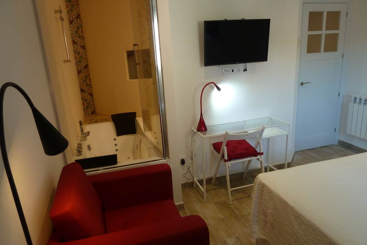 Habitación de hotel con una silla roja, un escritorio y una silla, una cama, un televisor montado en la pared y un baño con ducha e inodoro.