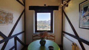 A dining area with a table set for two, a vase with dried flowers, and a window with a mountain view. - Casa Rural La Cortina (Cabrales)
