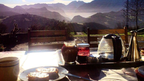 A table set with a plate of food, a cup, a jar, a kettle, and a thermos, overlooking a mountainous landscape. - Casa Rural La Cortina (Cabrales)