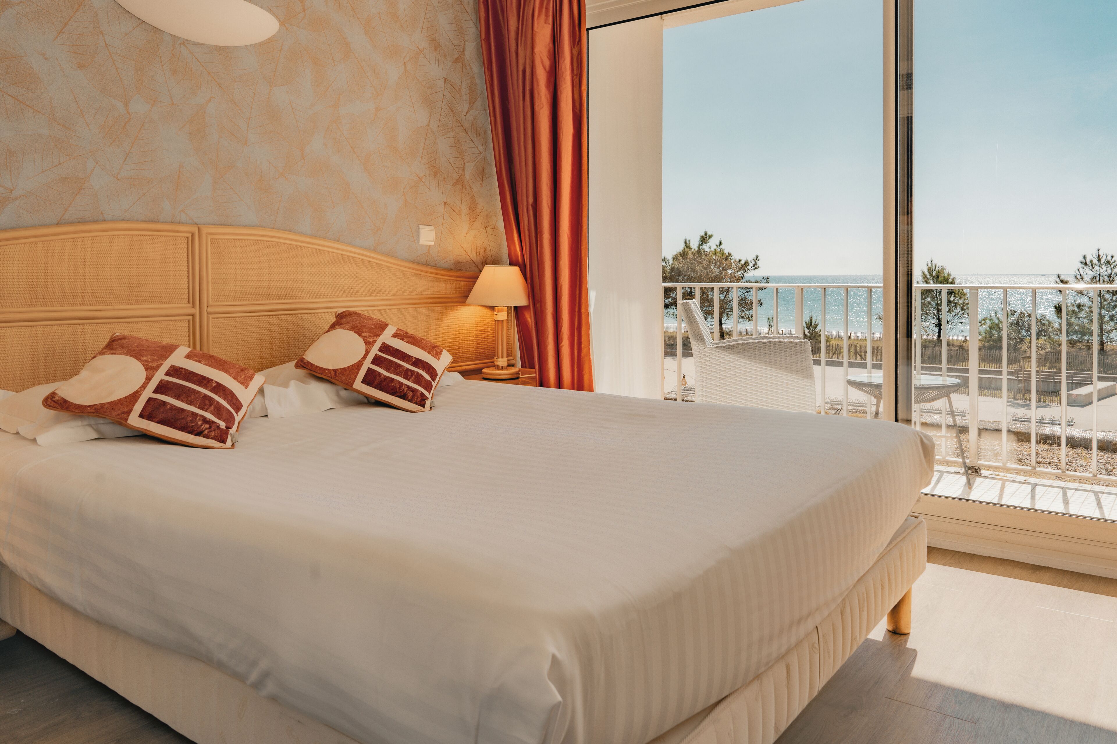 A hotel room with a large bed, a nightstand, a lamp, and a balcony with a view of the sea.