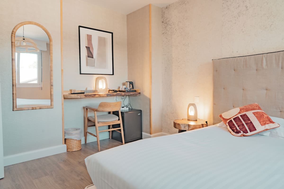A bedroom with a bed, a desk with a chair, a small refrigerator, and a wall-mounted shelf with a framed picture and a lamp.