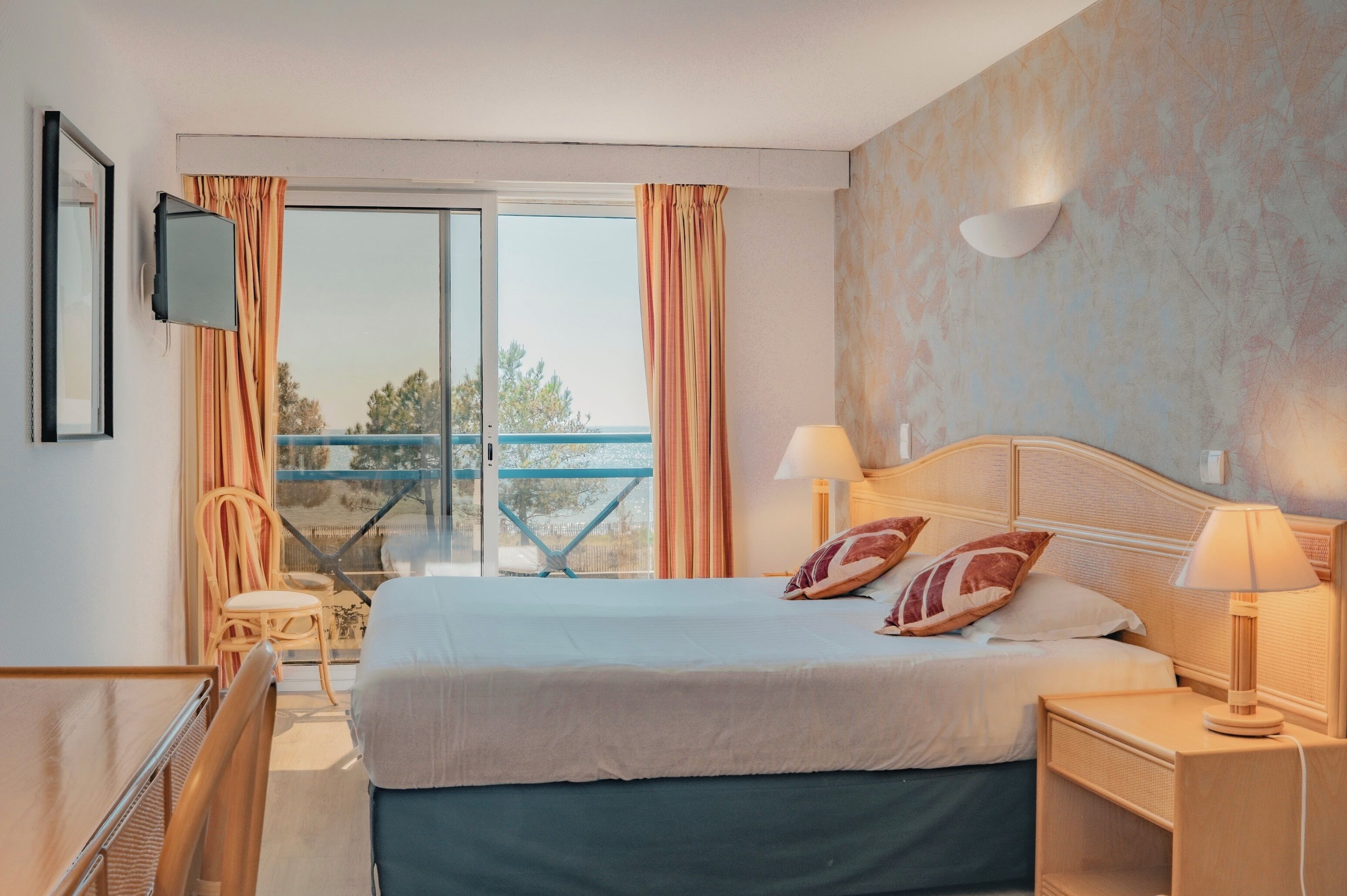 A hotel room with a bed, two bedside lamps, a TV, a chair, a balcony with a view, and a table.