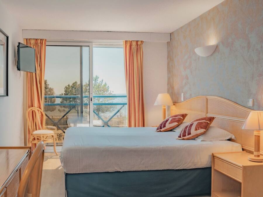 A hotel room with a bed, two bedside lamps, a TV, a chair, a balcony with a view, and a table.