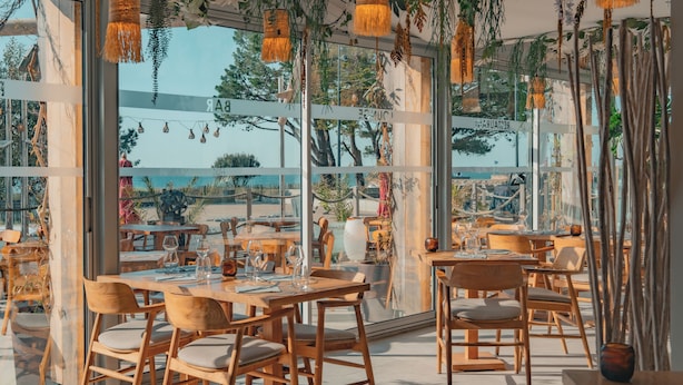 An indoor dining area with wooden tables and chairs, large windows, and hanging decorations. - Hôtel Edenia (Carnac)
