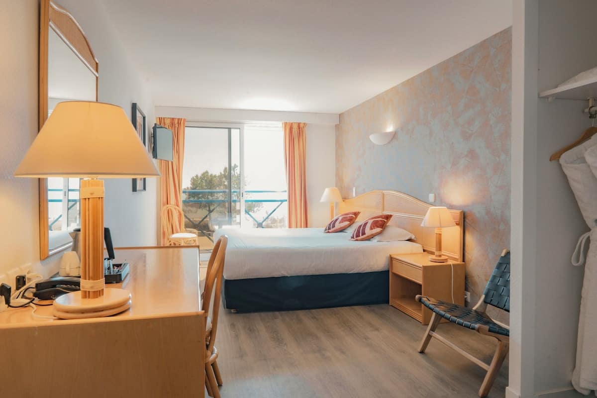 A hotel room with a bed, bedside tables, a desk, a chair, a lamp, a mirror, and a view of the outdoors.