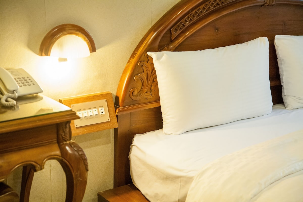 Een net opgemaakt bed met witte lakens, een houten nachtkastje met een telefoon, en een wandlamp.