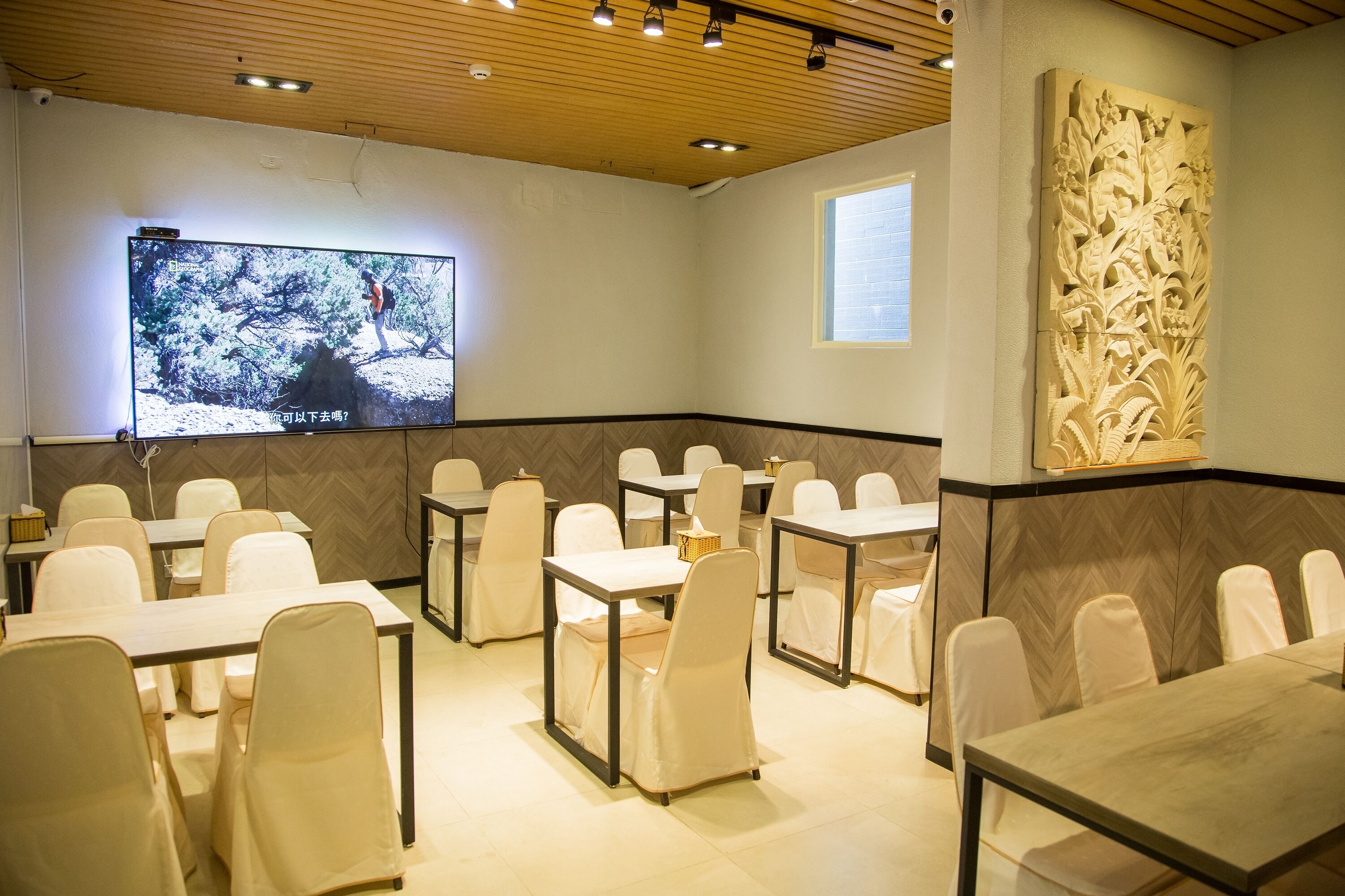 A conference room with multiple tables and chairs, a large screen displaying a nature scene, and a decorative wall panel.