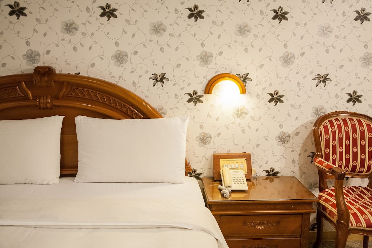 Een netjes opgemaakt bed met witte lakens, een houten nachtkastje met een telefoon, en een stoel met rode en gouden strepen.