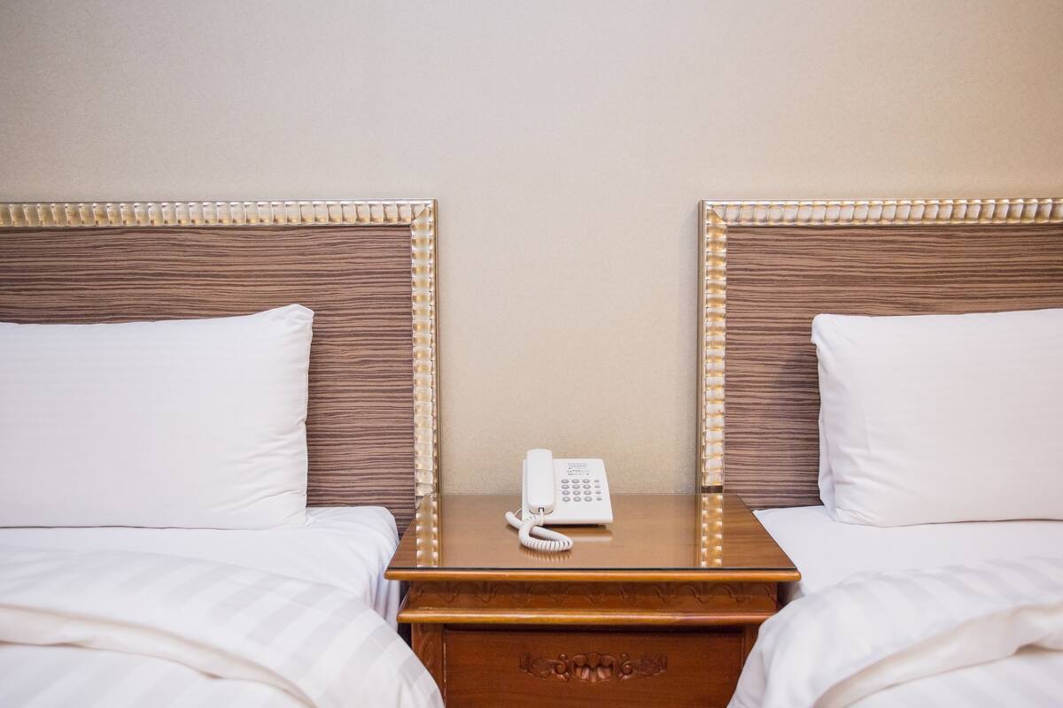 Een hotelkamer met twee bedden, elk voorzien van een houten hoofdbord en witte beddengoed. Tussen de bedden staat een houten nachtkastje met een witte telefoon erop.
