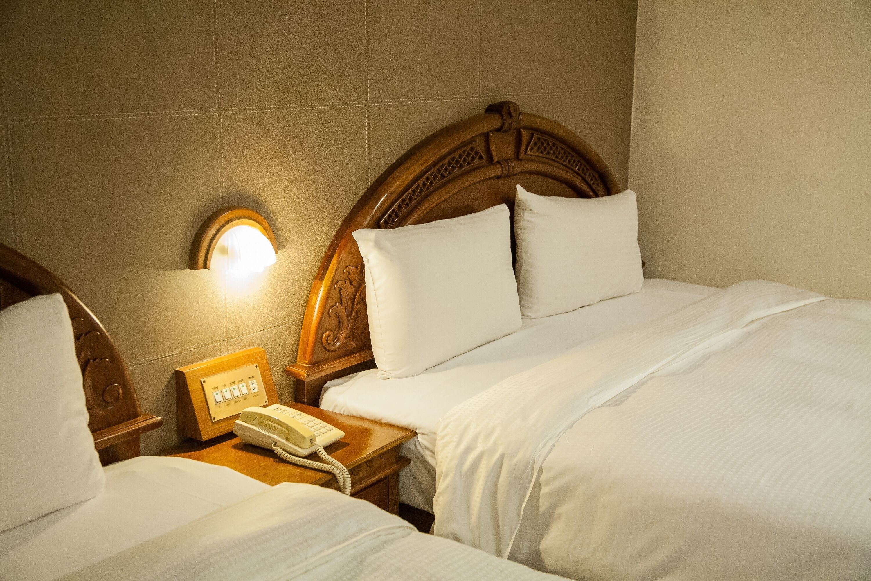 A hotel room with two beds, a wooden headboard, a bedside table with a telephone, and a wall-mounted light fixture.