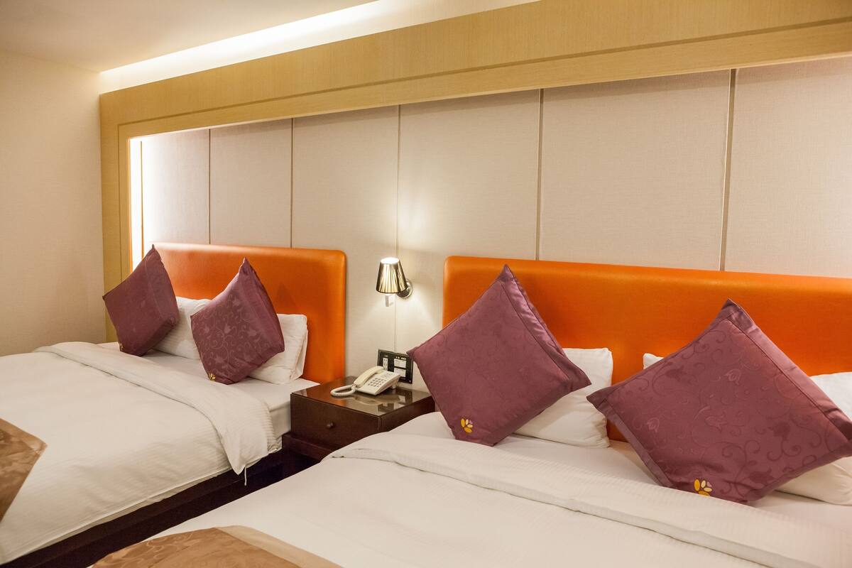 Een hotelkamer met twee bedden, elk voorzien van witte beddengoed en oranje hoofdeinden. Er staat een nachtkastje met een lamp en een telefoon.