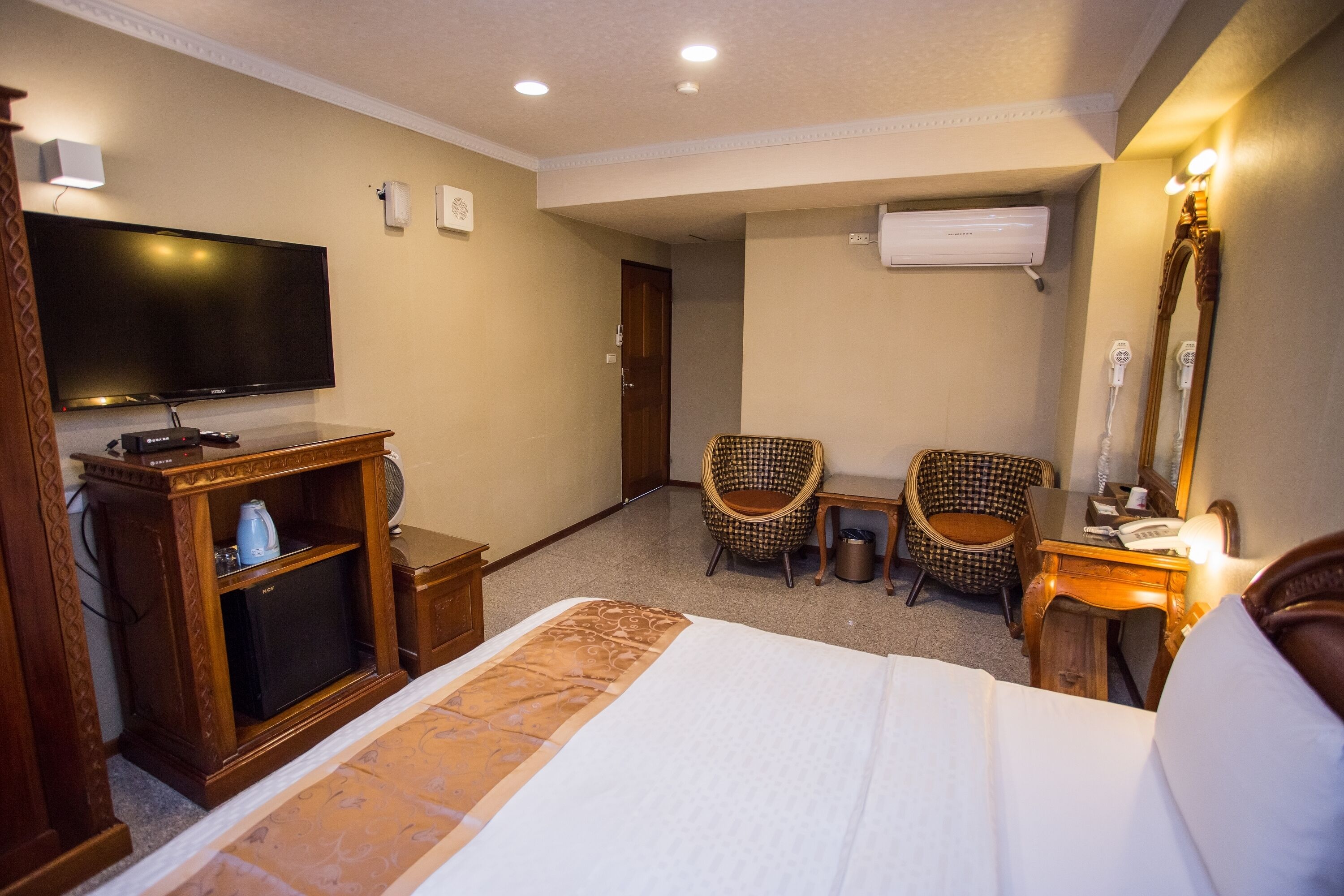 Habitación de hotel con una cama grande, un televisor de pantalla plana, un aparador de madera, dos sillas de mimbre, una mesita pequeña y un espejo.