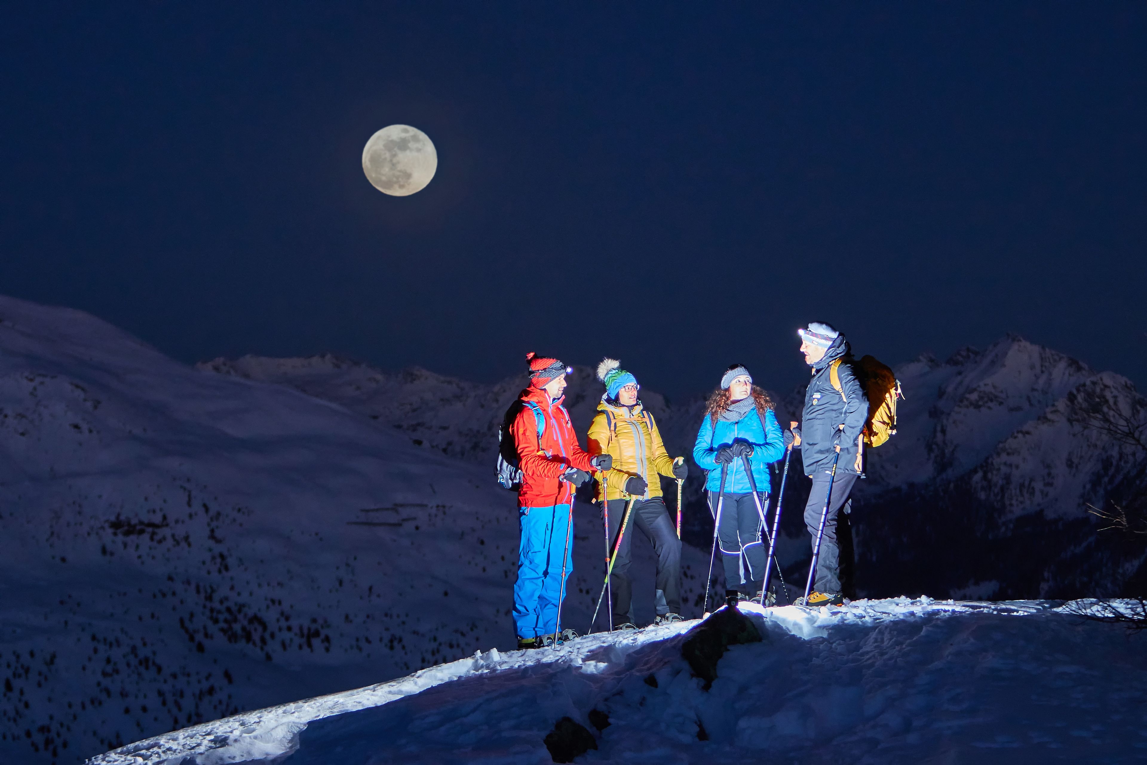 A group of people standing on a snowy mountain peak with a full moon in the background.