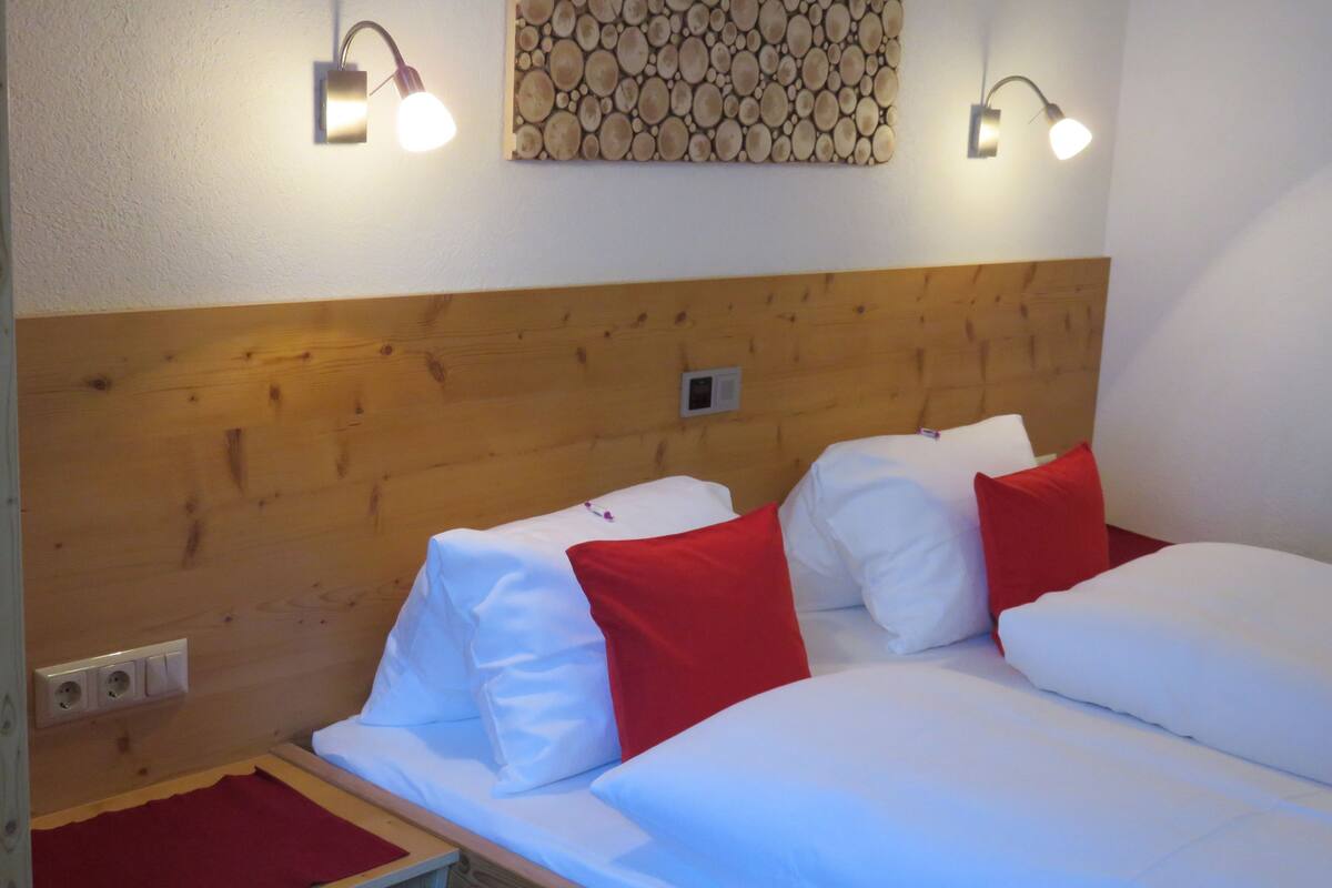 Quarto de hotel com cabeceira de madeira, duas camas com roupa de cama branca e vermelha, um cobertor vermelho na mesa de cabeceira e luminárias fixadas na parede.