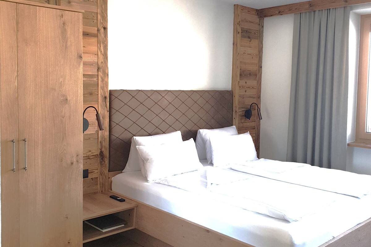 Uma cama de madeira com roupa de cama branca, uma pequena mesa de cabeceira e um guarda-roupa com espelho.