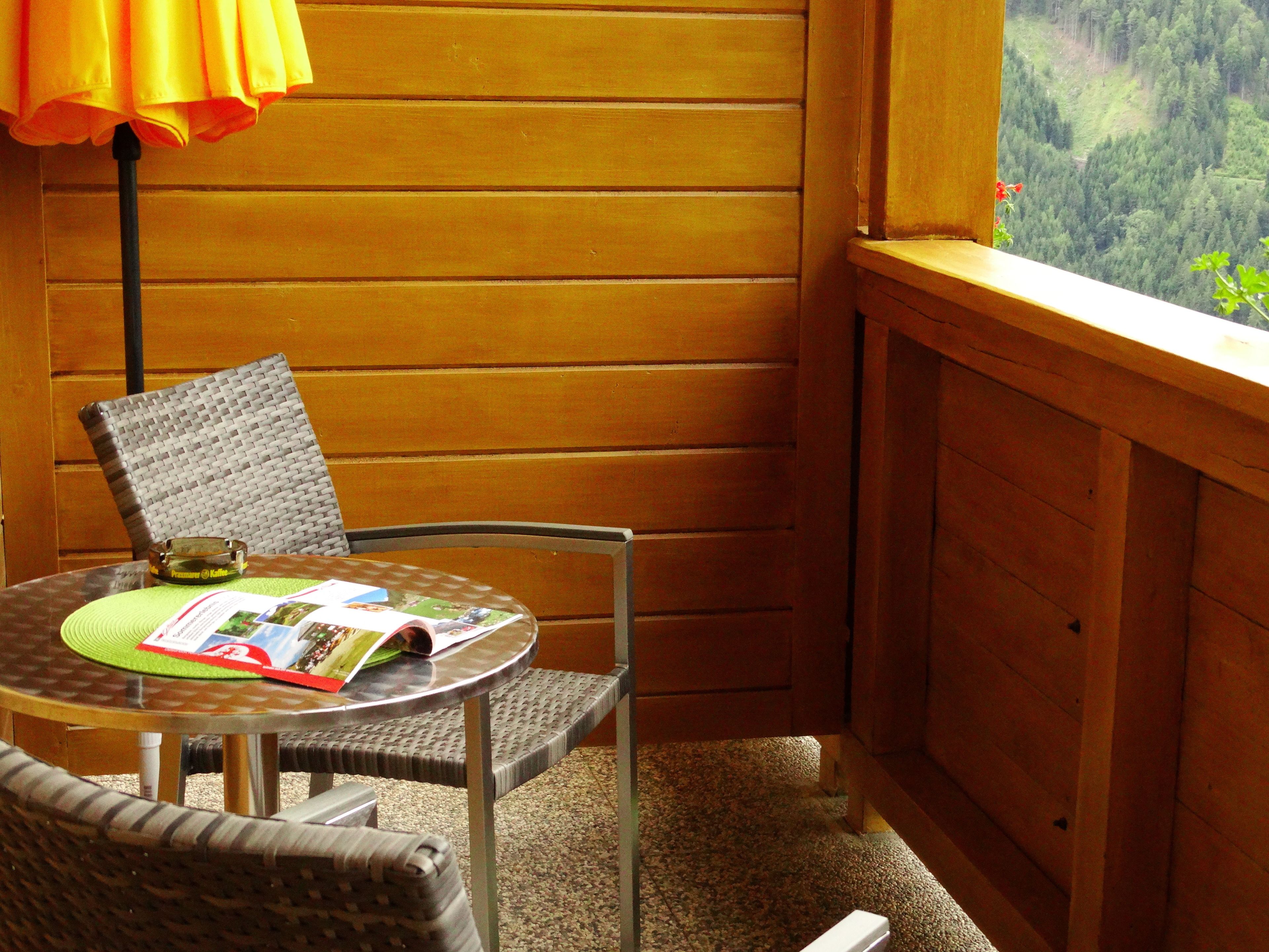 A corner room with a wooden wall, a table with a magazine, a chair, and a view of a forest through a window.
