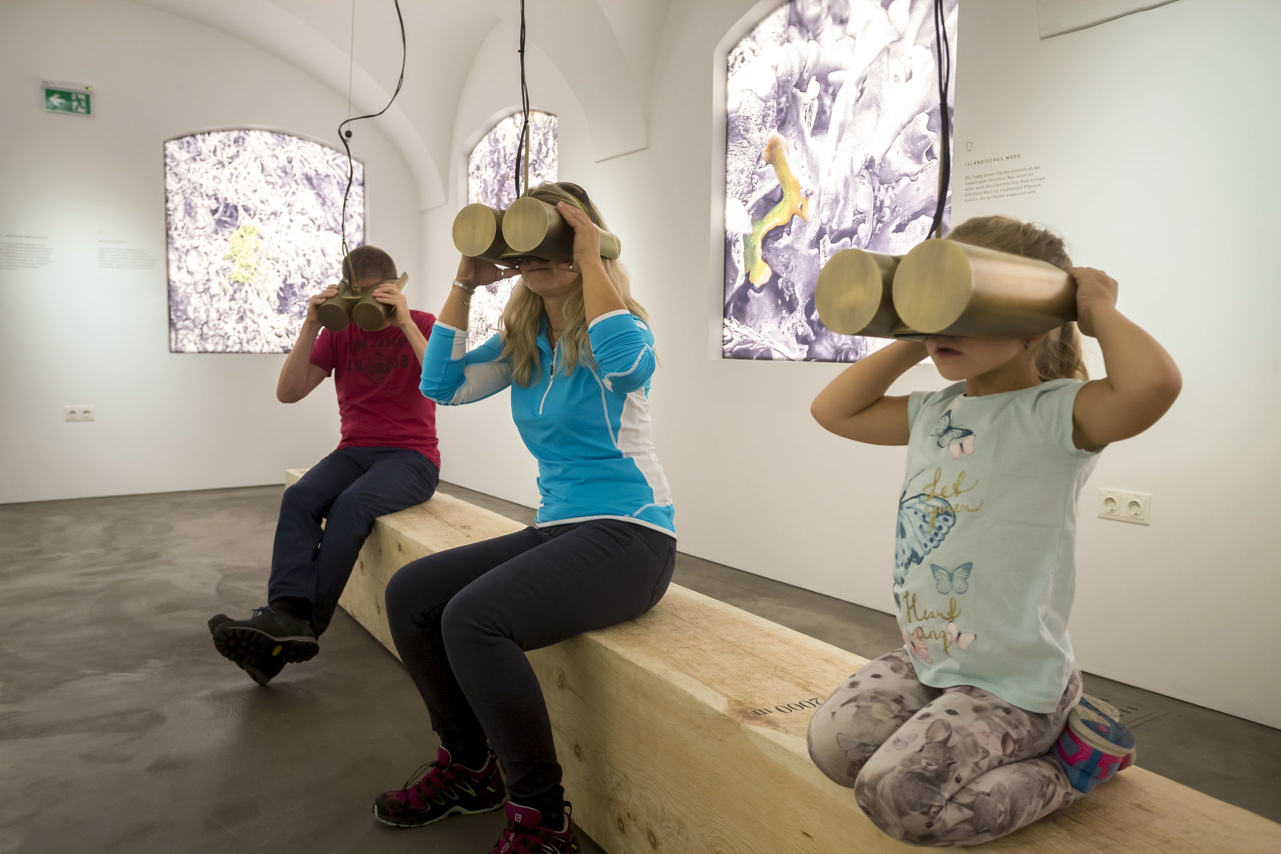 Three people in an art gallery with abstract paintings on the walls, wooden benches, and a concrete floor.