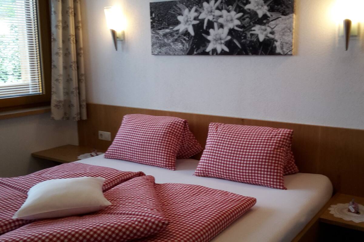 Quarto de hotel com cama coberta por roupa de cama xadrez vermelha e branca, um travesseiro branco e um quadro de flores fixado na parede.