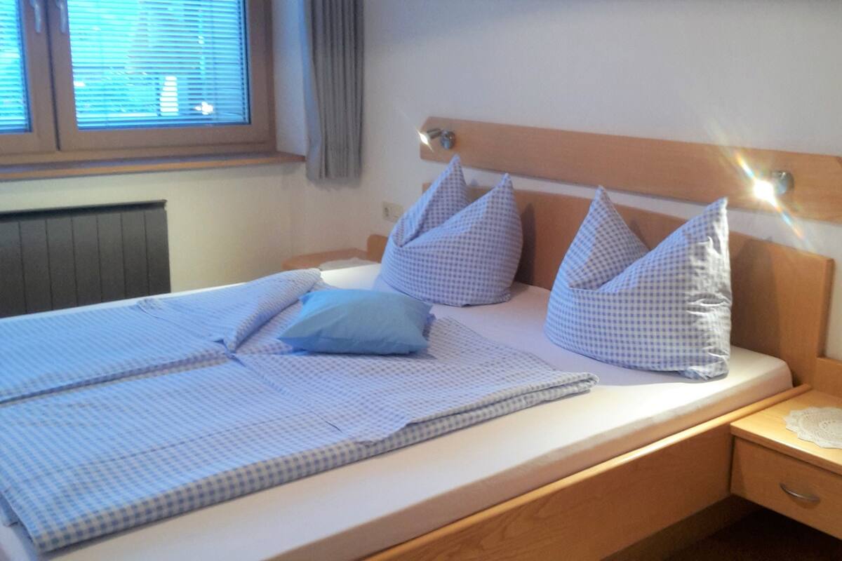 Quarto com cama, almofadas, mesa de cabeceira e janela com persianas.
