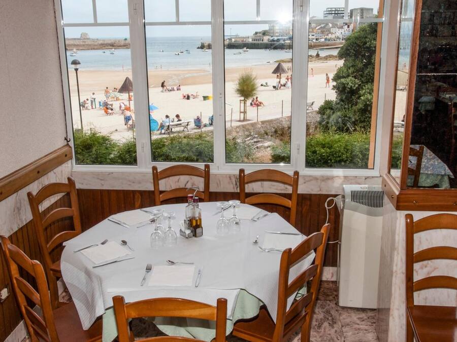 Un comedor con una mesa dispuesta para cuatro personas, con vistas a una playa con gente y sombrillas.