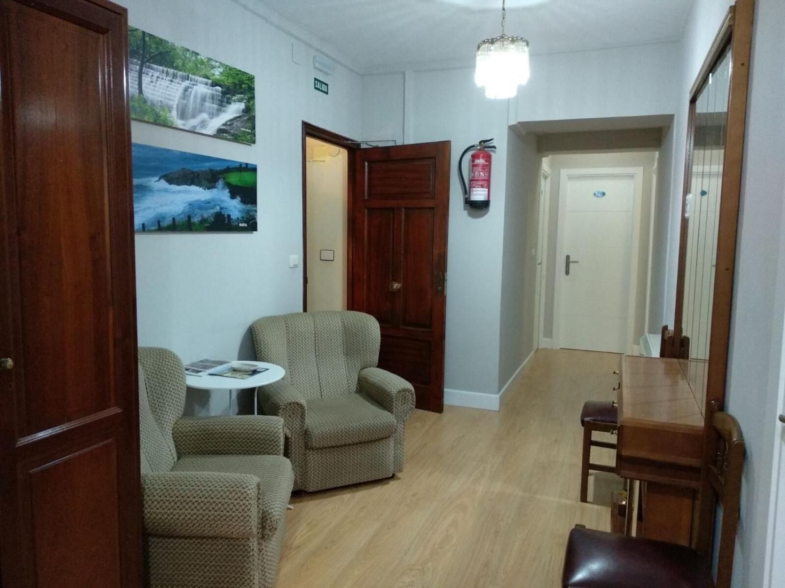 Un pasillo con piso de madera, una pequeña mesa redonda con una silla, un sofá y un armario de madera. Hay dos puertas, una abierta y otra cerrada, y un extintor de incendios montado en la pared.