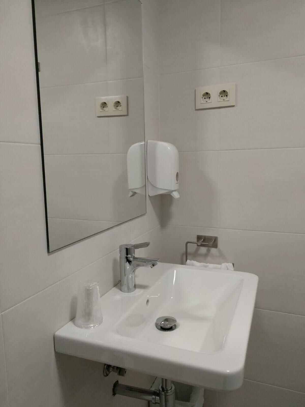 Un baño con lavamanos blanco, un espejo y dos dispensadores de jabón fijados a la pared.